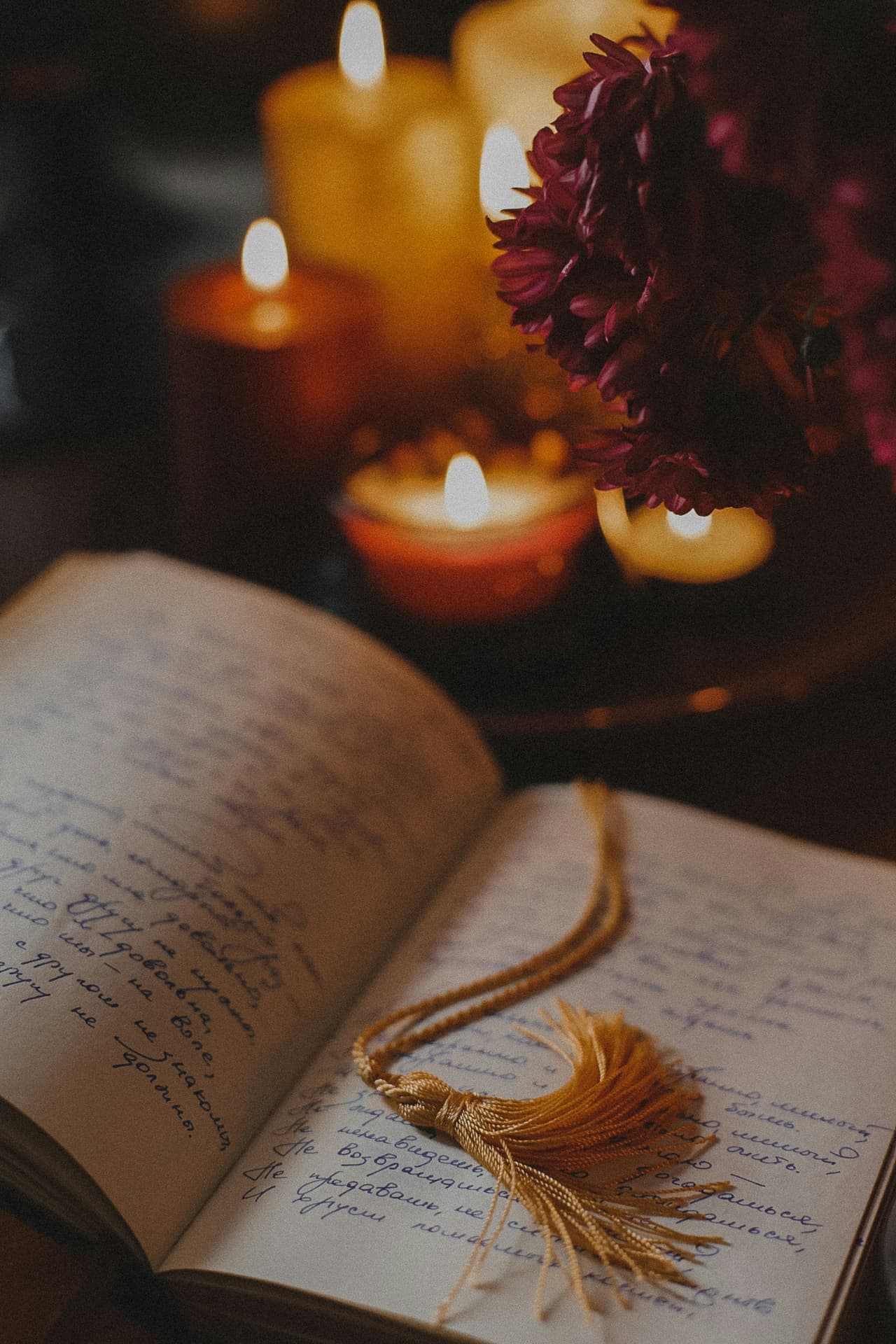 An open handwritten journal with a gold tassel bookmark, lit candles and flowers